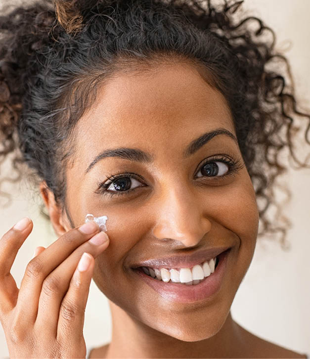 Smiling african girl with curly hair applying facial moisturizer while holding jar and looking at camera. Portrait of young black woman applying cream on her face isolated on beige background. Close up of happy attractive beauty woman caring of her skin standing on light brown wall with copy space.